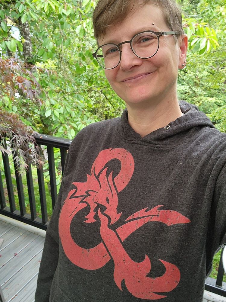 Me, a 53-year-old agender person, standing on my deck wearing a goofy grin and a hoodie with the Dungeons and Dragons logo.
