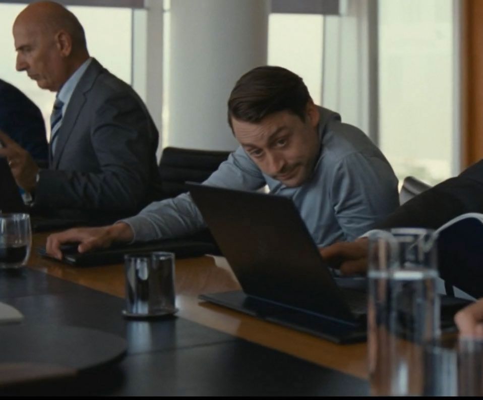 Scene from Succession where Roman looks sheepish in the board room after sending an embarrassing picture to his father