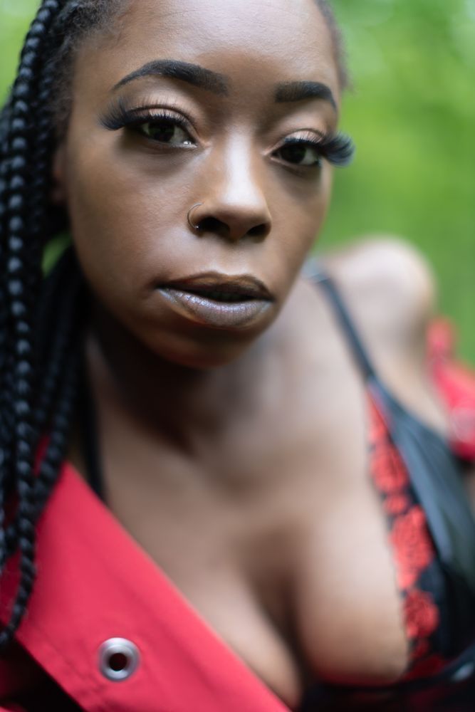 This close-up portrait burns with intimacy and erotic presence. A beautiful Black woman stares directly into the lens, her deep brown eyes framed by lush lashes and brows sculpted in perfect defiance. Her nose ring glints in the soft forest light, hinting at wildness restrained. The camera lingers at her chest, revealing the lace-trimmed edge of a red and black lingerie piece barely covered by a red jacket that hangs off one shoulder. Her full lips are parted slightly, the tension between vulnerability and strength playing across her face. There’s a smoldering energy here—an unapologetic, sensual confrontation that speaks volumes without a word. This is the face of erotic maturity, poised between mystery and invitation.
#NSFWArtPhotography #CloseUpErotica #BlackLingerieMuse #BoldAndBareFaced #EroticPortraits #Over40Seduction #RedAndBlackMagic #SensualGaze #AltBoudoirBeauty