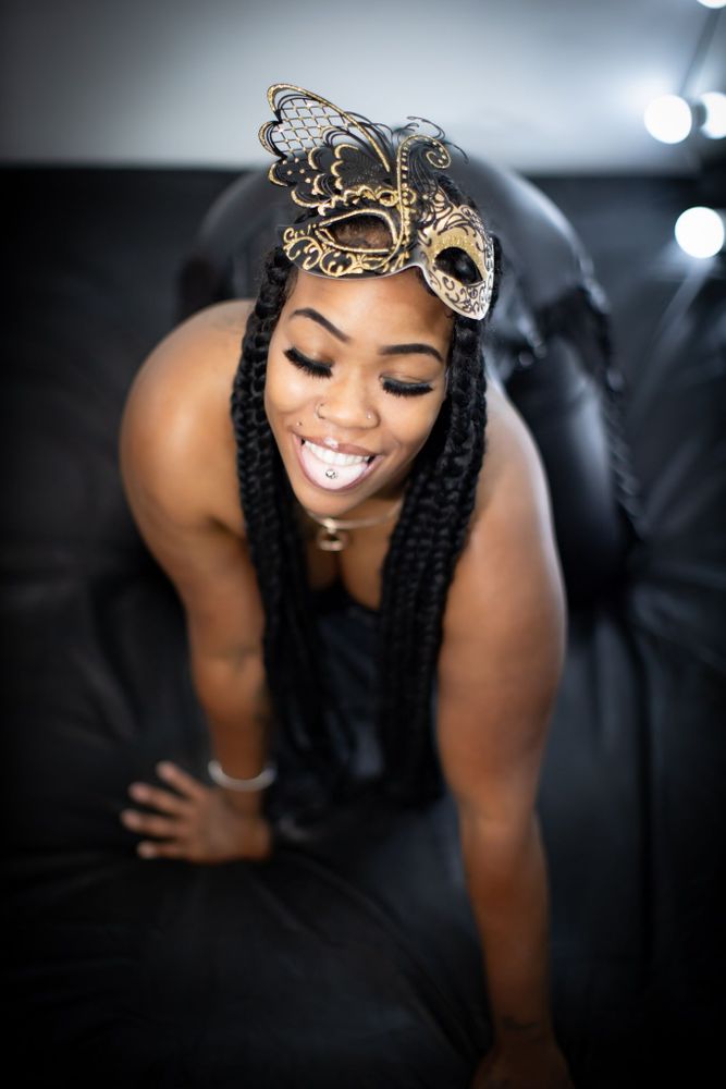 Shot from above, this captivating boudoir portrait showcases a confident woman perched low on a black-draped bed. Her long braids cascade over her shoulders, framing her glowing skin and sultry neckline. A metallic butterfly mask perches slightly raised on her forehead, revealing expressive eyes lined with lush lashes. Her smile radiates warmth and a hint of mischief as she gazes downward, dimples deepening with pleasure. A glint of silver from her tongue piercing peeks through parted lips. The body posture, low and feline, combined with the soft lighting and high contrast, makes her feel like a goddess just caught mid-purr. The elegant lighting setup enhances the curves of her shoulders and hips, while the shadows whisper secrets of desire. This photo speaks to a confident femininity, one that flirts with mystery while proudly owning her sensual presence.
#IntimatePortraiture #EroticEnergy #NSFWBoudoir #SensualMuse #BlackFemmeMagic #GlamErotica #MaskedDesire #SoftDommeVibes