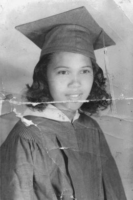 A young girl in cap and gown in a high school graduation photo in 1958. The black and white image has creases that suggest it has survived damage.
