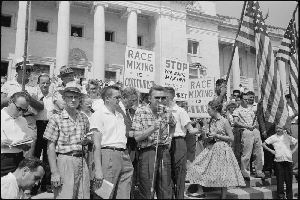 Black and white photograph of adults standing in front of a building with columns made by the photographer John T. Bledsoe on August 20, 1959. A man is speaking into a microphone. The group is protesting the admission of Black students to public high school. 