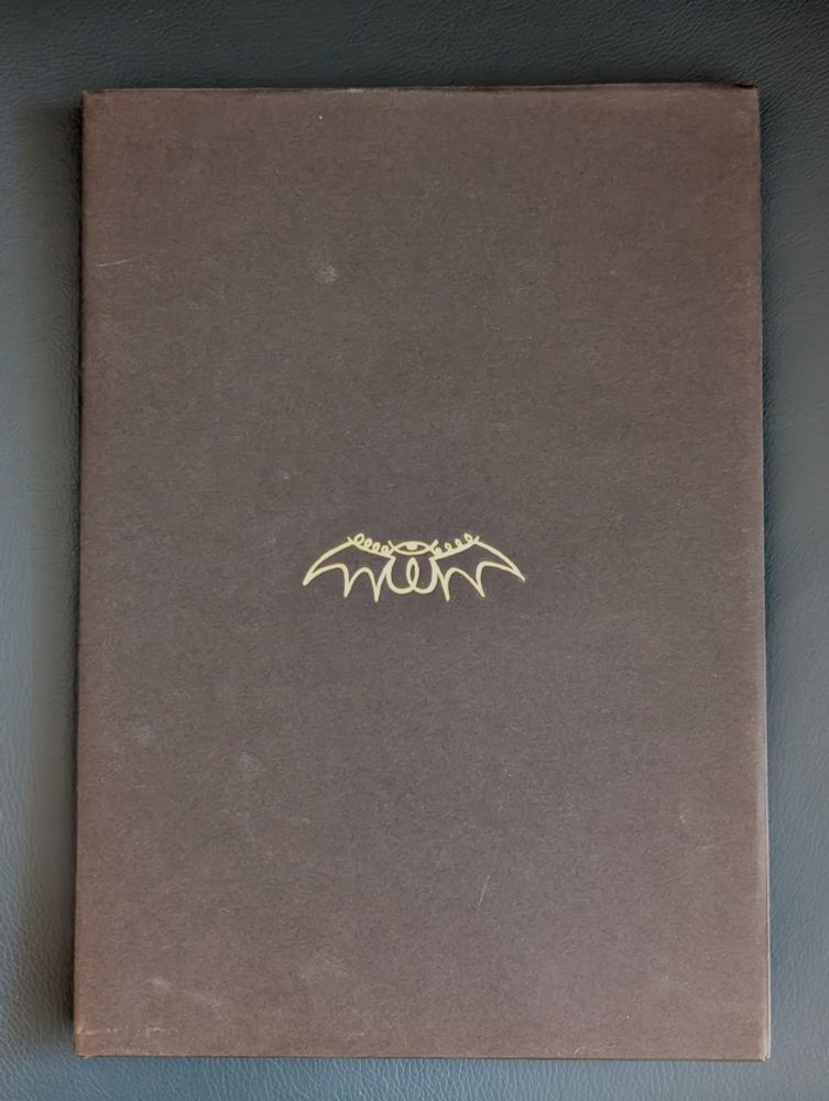 A black hardcover book with a minimalist design. The only visible marking is a small gold emblem in the center, resembling a stylized bat with decorative flourishes above its wings.

