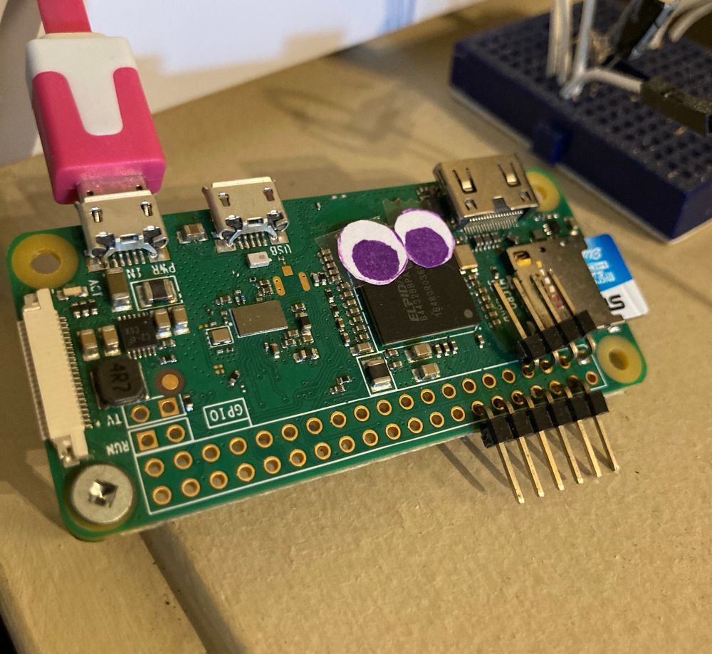 closeup of a pi zero with a pink usb cord and purple hand-drawn googly eyes on its cpu