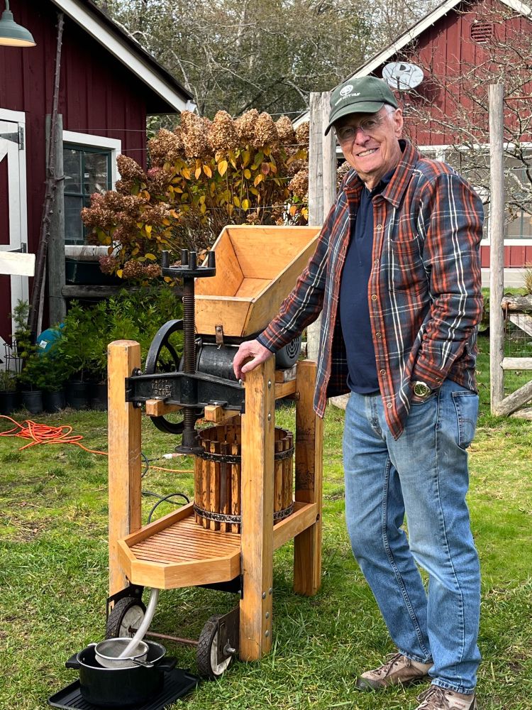 Photo of a man in a plaid flannel shirt outdoors next to a wooden hand cranked apple press.