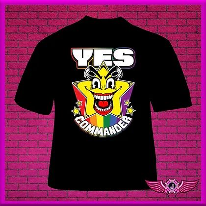 A black t-shirt on a pink brick background. The t-shirt features a drawing of a cartoon star with a manic grinning face and lipstick. The star is leaving a rainbow trail, and four smaller stars burst from the sides. The words "YES COMMANDER" frame the artwork, with YES in a bold straight line across the top, and "COMMANDER" in a downward arch across the bottom.