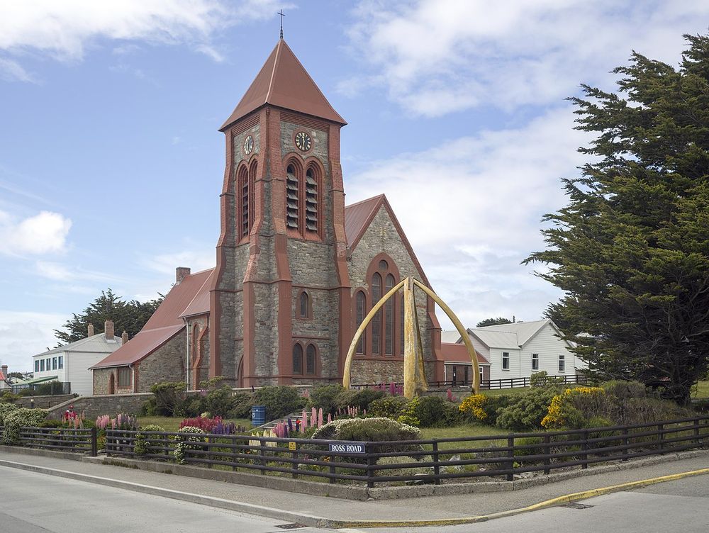 Modern photo of Christ Church Cathedral, Port Stanley, Falkland Islands. A Gothic Revival Church, this photo was taken in the summer and show flowers and gardens outside the church and blue skies and clouds behind it.