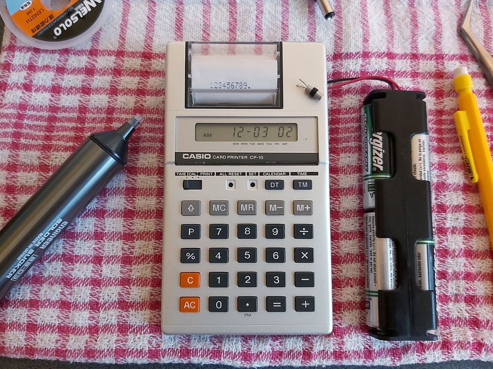Photo of a Casio Card Printer CP-10 calculator showing the time (12-03 02) on its screen and a printout of "123456789.". A battery pack of four AA cells is attached to the right of the calculator. A quartz crystal resonator is sitting on top of the calculator.