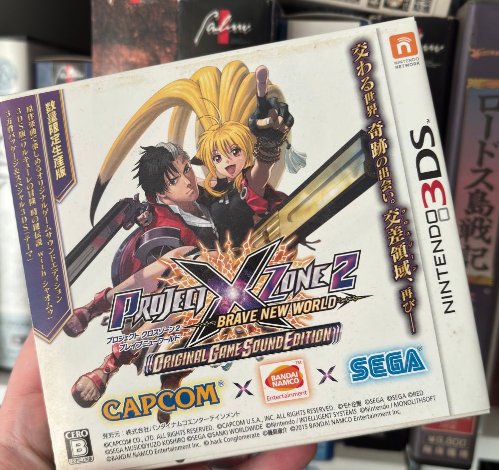 A photo showing the Japanese box for Project X Zone 2 (3DS) held up to the camera. The cover shows Chipper Lady cheerily raising an arm, while being held by Serious Guy. They have unrealistic guns in their free hands.