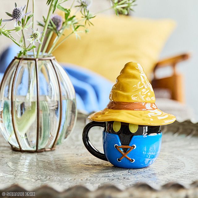 A photo showing a ceramic mug sitting on a table, next to a glass vase filled with flowers. The mug's shape is heavily inspired by Vivi, Final Fantasy 9's black mage character. It has a wide-brimmed ceramic pointy wizard's hat as a lid.