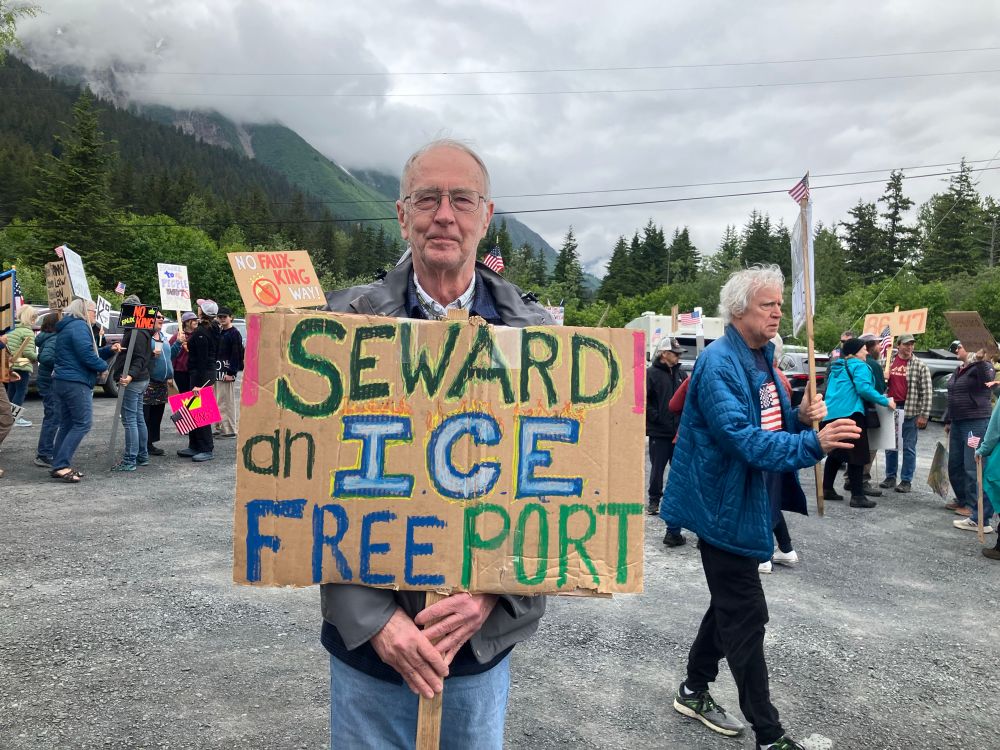 A man stands with a sign saying ‘Seward: an ICE free port’ with other signs and protesters in the background. Seward is one of the most northerly year-round ice-free ports in Alaska. We don’t like Immigration and Customs Enforcement either.
