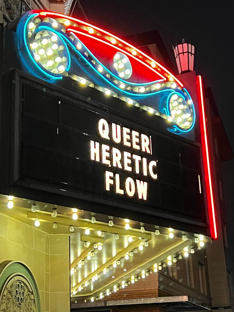 A theater marquee displaying the movie titles “Queer”, “Heretic”, and “Flow” on top of each other so it reads as “Queer heretic flow.”