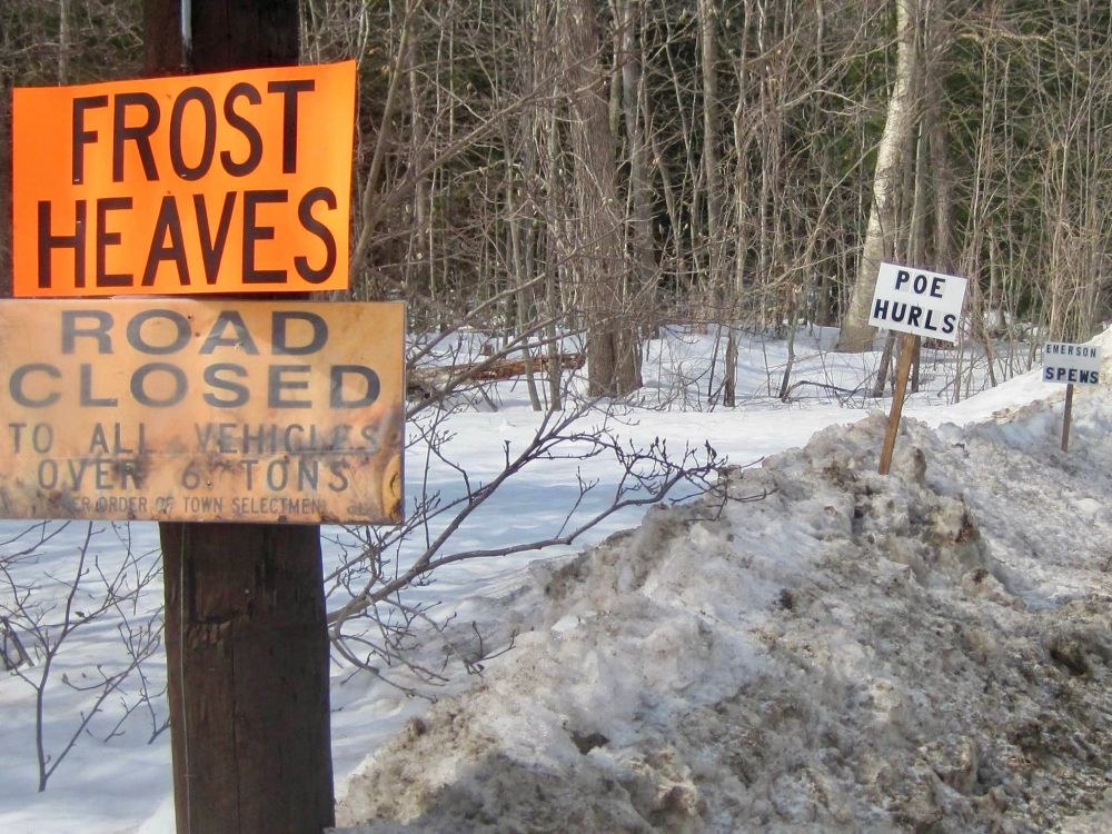 A snowy road w orange road sign that says “Frost Heaves” and Road Closed , with a handmade sign 10 yds down road that says “Poe Hurls” and another 10 yds further on “Emerson Spews”