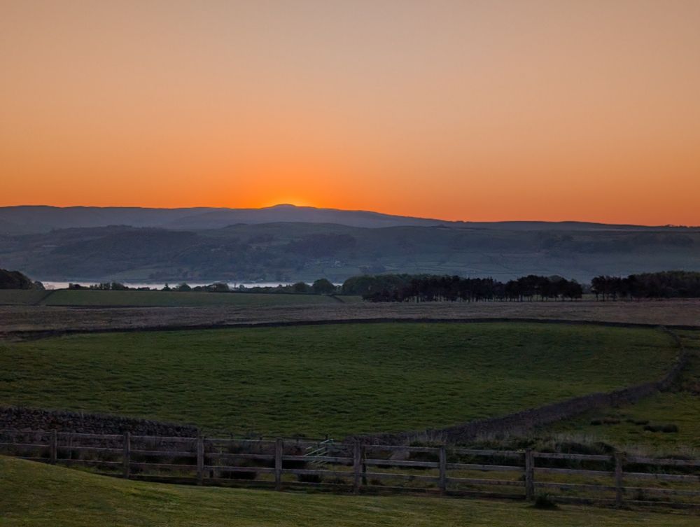 A view across some fields to distant hills. The sun is just rising behind the peak of the tallest hill, deep orange fading into the palest of blues. At the lowest point in front of the hills early mist mist lingers, looking like a silvery band of water in the distance. 