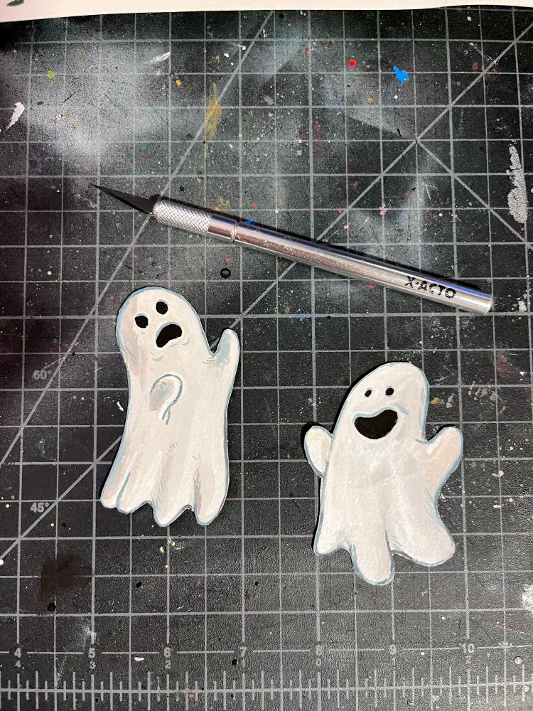 Two ghosts that are a mix of cut paper and painted textures. One of them is cheerful while the other is dismayed. They are on a self-healing cutting mat and next to them is an x-acto knife. 