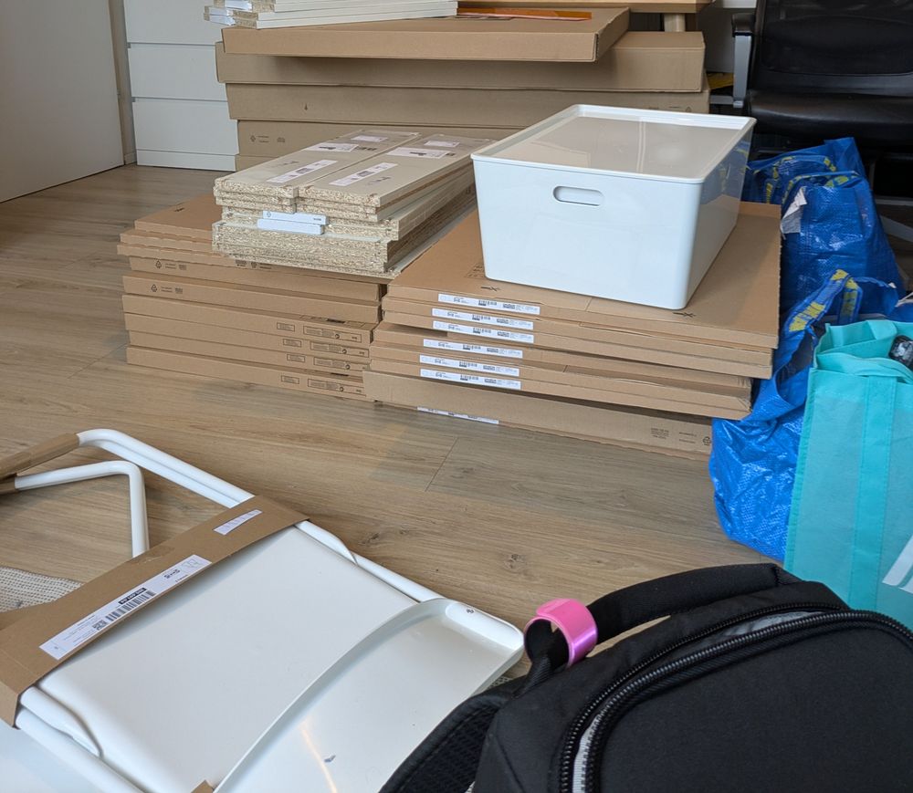 A huge pile of Ikea packages stacked on each other. A white chair and a black backpack in the front.