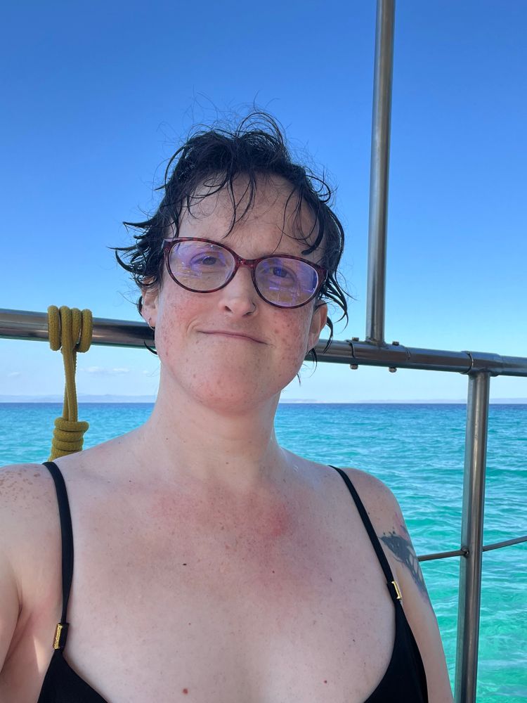 A white woman with pixie cut length brown hair and brown eyes she is wearing brown glasses and a black tankini. She is sitting on a boat with the sea behind her.