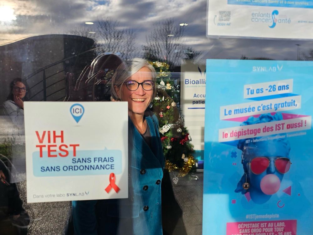 Clara de Bort est derrière la vitre du laboratoire d’analyse Medicale sur laquelle sont affichées des informations sur le fait que ce laboratoire propose le test VIH sans frais et sans ordonnance, et qu’il propose aussi le test de 4 autres infections sexuellement transmissibles (pour les - de 26 ans, c’est 4 autres tests sont gratuits)