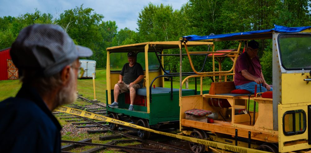 A small and open train-like vehicle riding the train tracks with an older man watching as they pass
