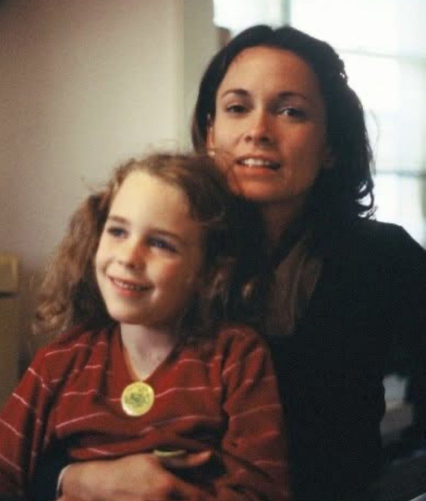 A dark haired woman looks into the camera. A young girl with a red sweater is sitting in her lap. 