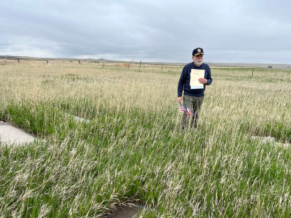 A man with a baseball hat and white beard holding a manilla folder stands in the high grass of an overgrown cemetery 
