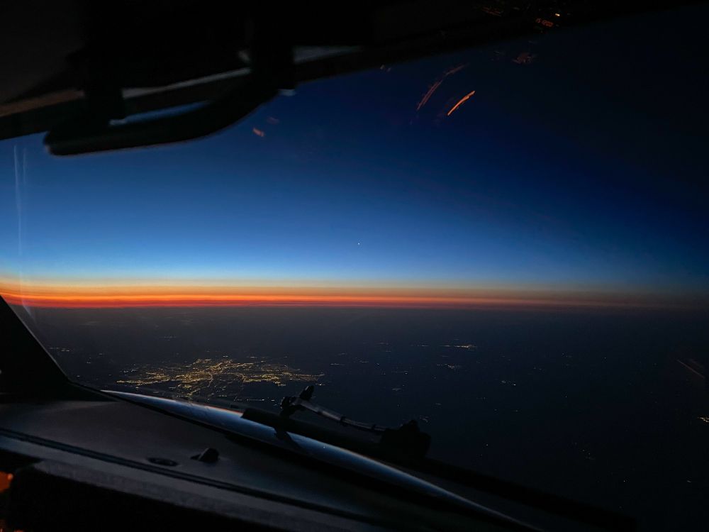 View from an airplane cockpit heading eastbound at high altitude during sunrise. The sky transitions from deep blue at the top to a vivid orange and red band near the horizon. City lights are visible on the ground below.