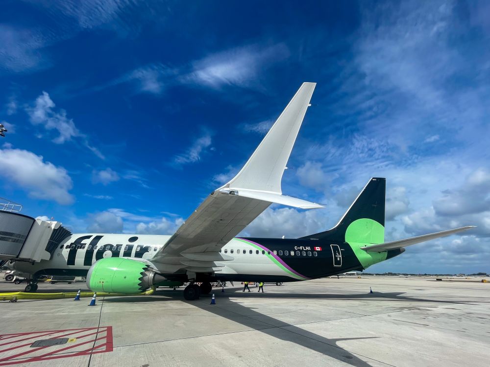A Flair Airlines 737 MAX 8 parked at the gate on a sunny day, with a bright blue sky and scattered clouds.