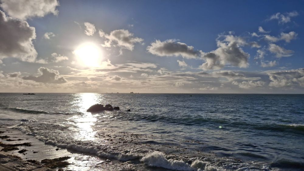 a colour photo of a beach with the tide coming in covering the sand, with white clouds on the horizon with rocks in the foreground on the left