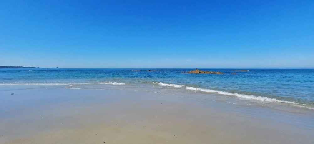 a colour photo of a beach with a calm receding tide, blue sea and blue sky