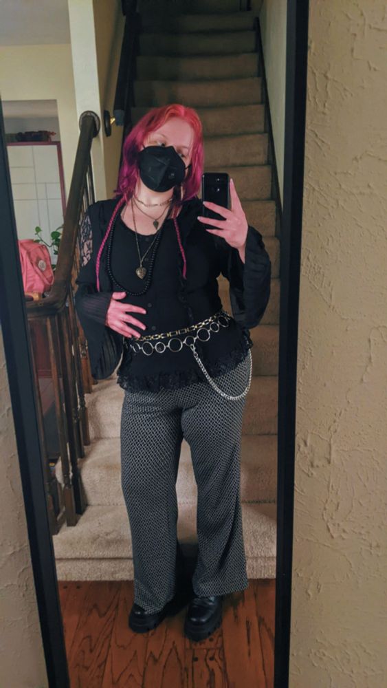 Mirror selfie of Jules, a white nonbinary person with shoulder length pink and purple hair with long braids on either side. They're wearing black pants with a cream diamond print like sparkles, a black shirt with ruffles and lacy bits at the shoulders and along the bottom, layered chain belts, layered necklaces, black platform boots, and a black face mask.