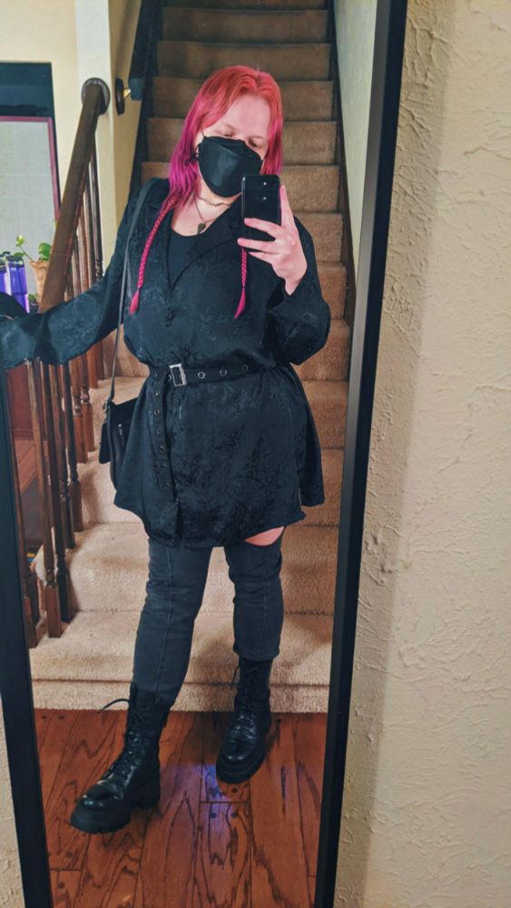 Mirror selfie of Jules, a white nonbinary person with medium length purple and pink hair with two longer braids on either side. They're wearing an oversized silky black button down with a subtle baroque pattern sinched at the waist with a black single grommet belt with a rectangular silver buckle, dark grey skinny jeans with cutouts at the thighs barely peaking out from under the shirt, layered silver necklaces, big black leather boots, and a leather bag over their right shoulder. They're standing with their right arm extended and resting on the bannister of stairs behind them, hips cocked to the left, and with their right leg kicked out in front of them 