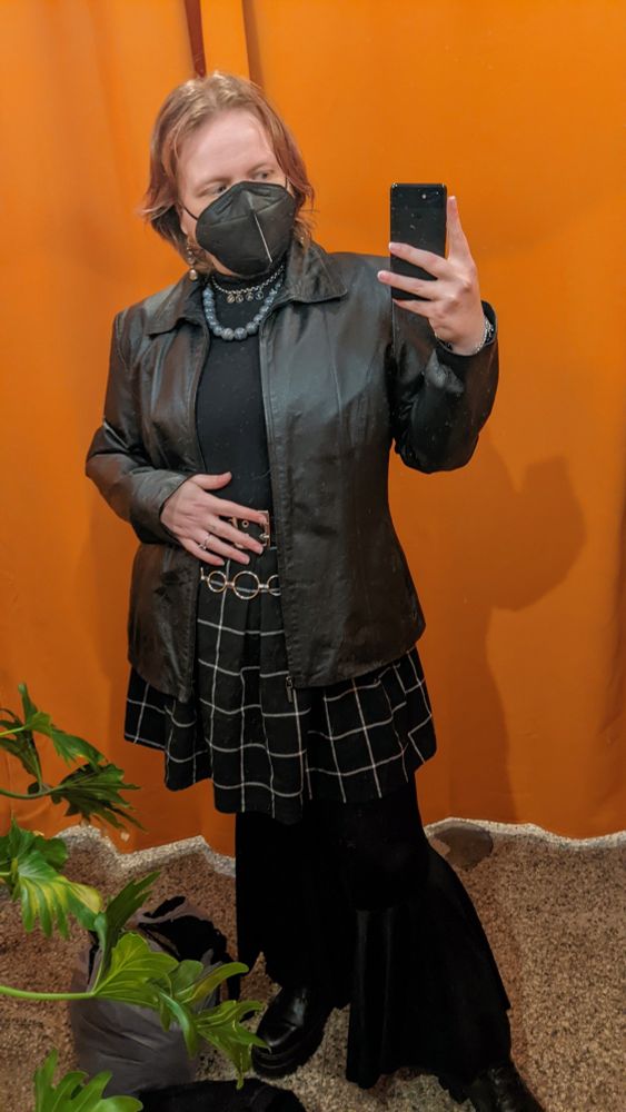 Selfie of Jules in a dressing room mirror with a bit orange curtain behind them, wearing a well fitting leather jacket