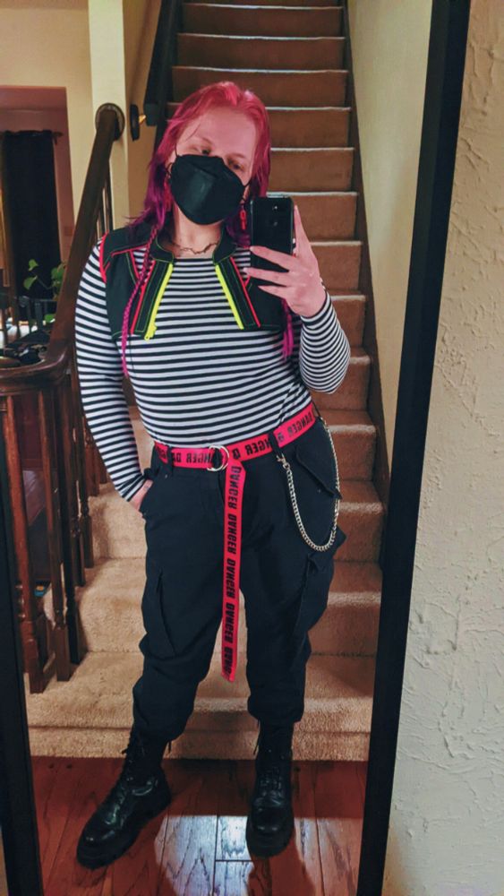 Mirror selfie of julian, a white non-binary person with medium length purple and pink hair with two longer braids in each side. They're wearing black cargo pants, a long sleeve black and white horizontal striped shirt, a cropped dark grey vest with red trim and a yellow zipper, a red belt that has a long tail in the front and reads "DANGER" in black letters repeating along the entire length of it, black platform boots, a silver belt chain, red acrylic chain earrings, and a black face mask. They're standing with their left hand in their pocket, hip cocked and head tilted to the right.