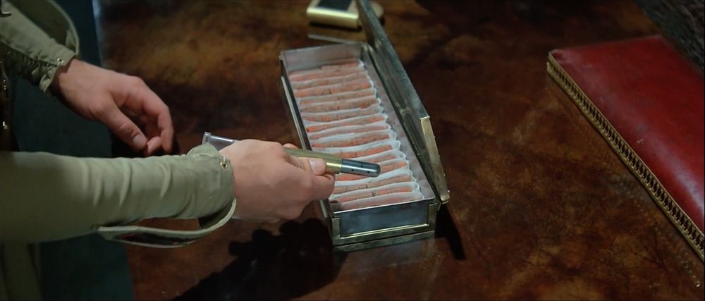 A still from Dune (1984) showing Paul Atreides in his room on Arrakis, wearing khaki green military sleeves, using a handheld scanner to check an open metal case containing rows of bright orange spice samples in individual sections. The scene takes place on a dark wooden table with rich upholstery and ornate details visible around, emphasizing the scientific examination of the precious melange.