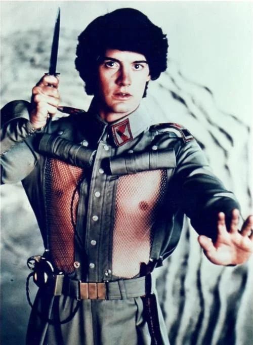 A still from the 1984 film Dune showing Kyle MacLachlan as Paul Atreides in House Atreides military uniform. He holds a knife in his raised right hand while extending his left hand forward. The costume features distinctive circular mesh panels exposing his chest/nipple area, set against metallic armor plating. His expression is intense and heroic despite the uniform design choices.