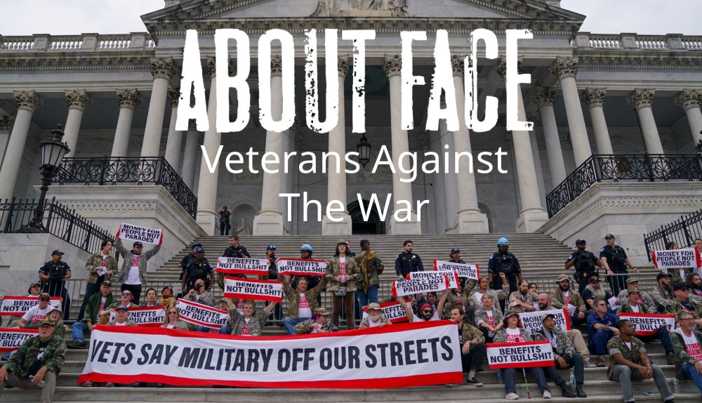 Veterans protest on the steps of a neoclassical government building with white columns. Large group of protesters, many in military uniforms or veteran attire, hold red and white signs reading 