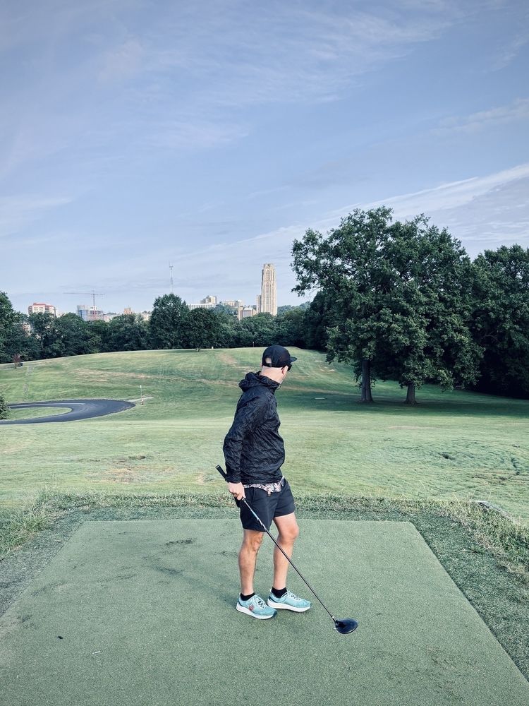 A person holding a golf club stands on a golf course with trees and a cityscape in the background.