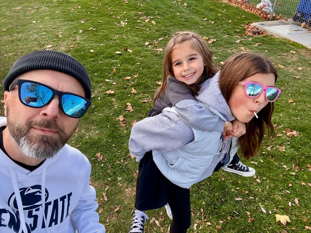 A man in sunglasses and a beanie takes a selfie with two smiling girls, one giving the other a piggyback ride, on a grassy area with scattered leaves.
