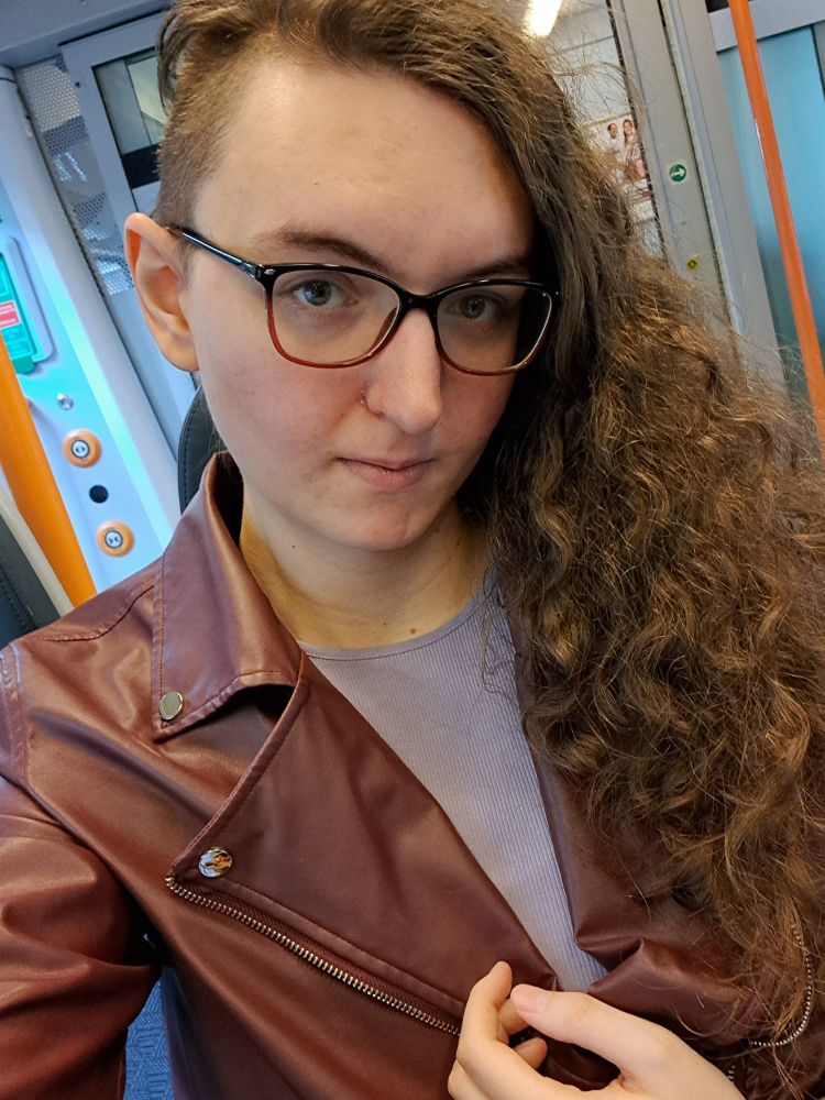 A picture of rin, a young pale woman with curly hair and the right side shaven in a burgundy leather biker jacket, a lavender top, and a red-brown pair of glasses. The interior of a train is visible in the background.