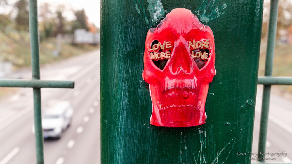 
Positive Skull Says Love More, More Love 

Skidmore Street I-5 Overpass, Portland, Oregon

Horizontal Color Photograph: A red plastic skull with the eye sockets painted black is affixed to a green post on the bridge. To the viewer's left and right the widely spaced mesh of the fences along the bridge are visible, and in the background, I-5. In gold paint, the Love More is written on the right eye socket, and More Love on the left one.  To the viewer's left, a white vehicle is visible heading north on I-5. 