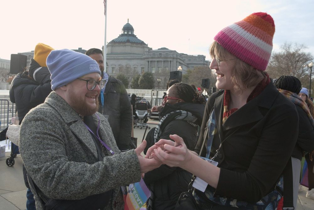 Valorie Van-Dieman from Assigned Media  in a hat with lesbian pride colors speaking excitedly with a trans man in a grey coat and baby blue hat who is participating in a protest in front of the Supreme Court in December 2024.