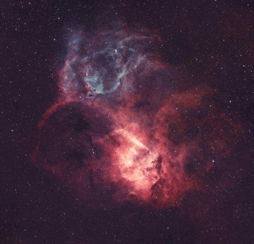 A bright nebula consisting of a light blue upper and light/dark red bottom areas. A background of stars shines within the nebula and in the background edges. 
