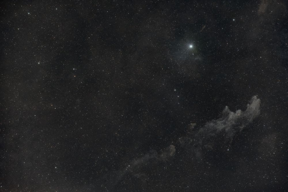 A field of stars with the grey dust of the witch head nebula to the bottom right and the bright star Rigel situated above it. 