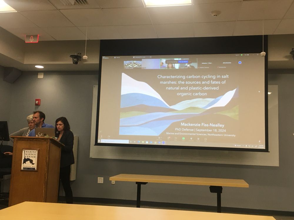 Standing in front of a podium with the title slide of my PhD defense presentation behind me, titled “Characterizing carbon cycling in salt marshes: the sources and dates of natural and plastic-derived organic carbon.”