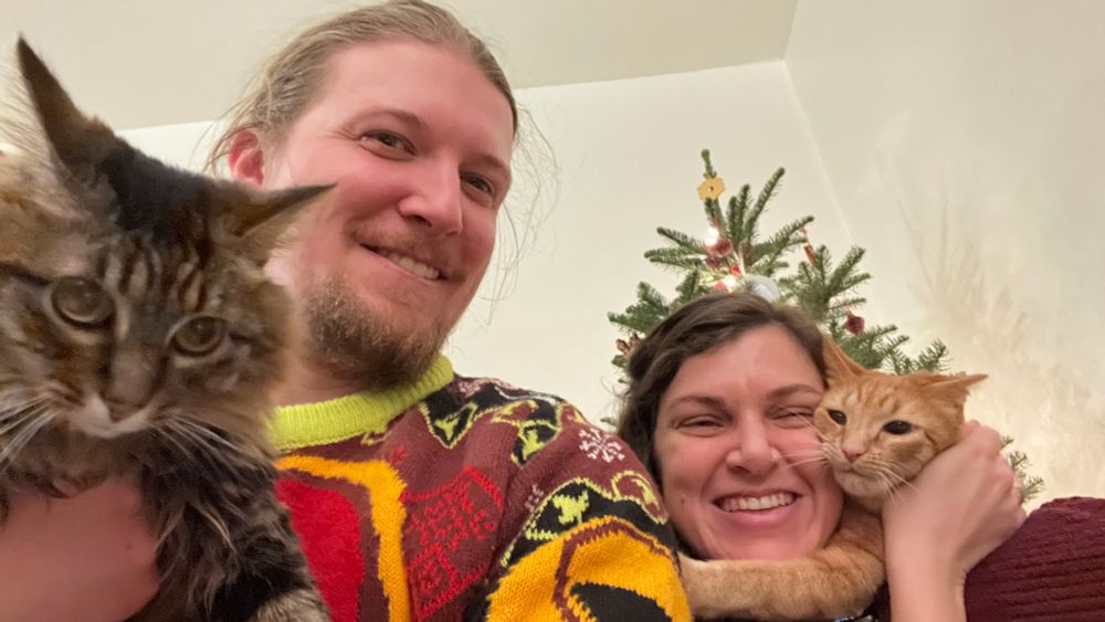 My husband and I are holding our two cats (who do not want to be held) in front of our Christmas tree