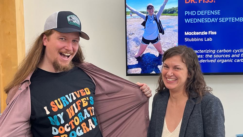My husband and I standing in front of a TV monitor that says “Congratulations, Dr. Fiss!” His shirt says “I survived my wife’s doctorate program.”