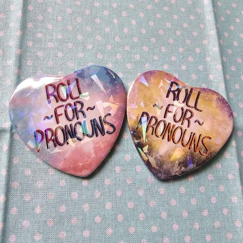 Two heart buttons with broken glass holo film, one themed after the transgender pride flag and the other nonbinary, that say, "Roll for Pronouns".