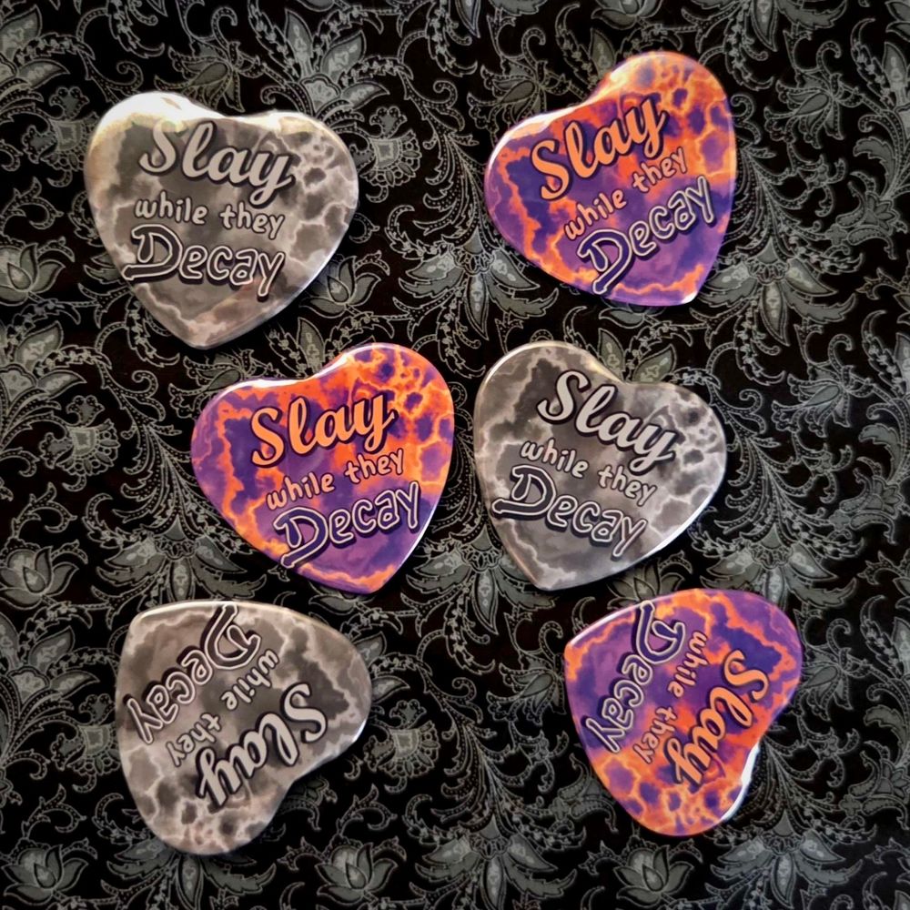Product photo of heart buttons scattered together, all saying "Slay while they Decay". There's two variants, one in orange and purple while the other is in black and white.