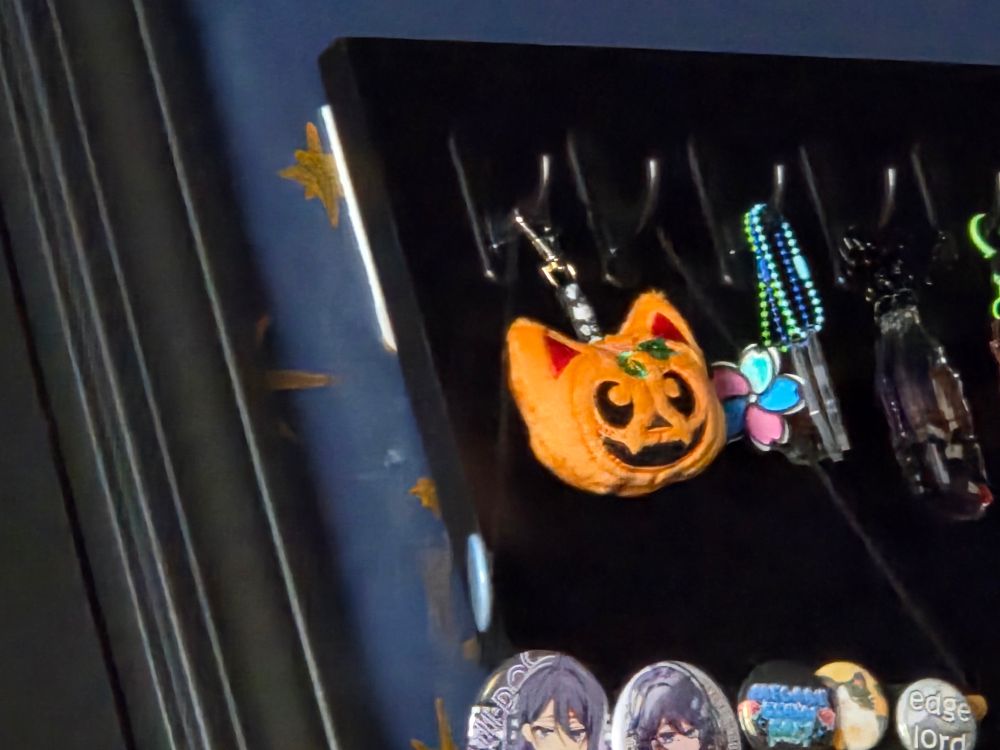 A plush keychain of a jack o' lantern shaped like a cat, hanging on my pegboard.