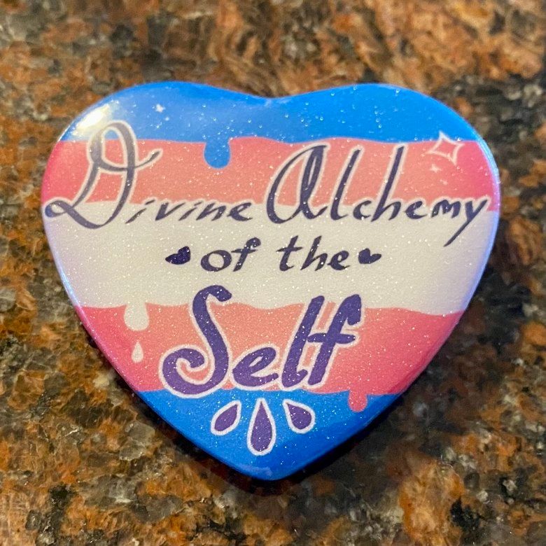 Heart button with sugar glitter holo film, themed to the transgender pride flag. Written is, "Divine Alchemy of the Self".